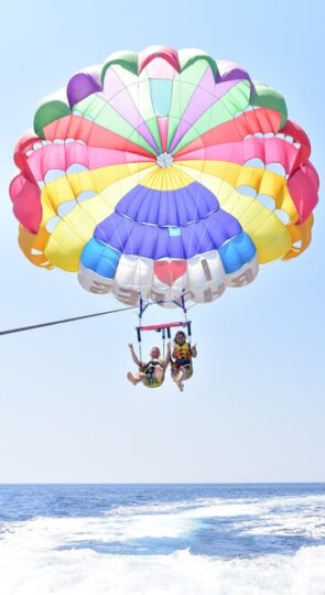 This is a picture of two people doind parasailing, the parasailing is colourfull and the people look happy above the sea at sotos watersports in rhodes
